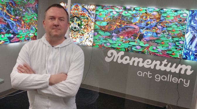 Artist matt corones stands in front of the Momentum West gallery his unique nostalgic, slightly chaotic mixed media art pieces hang on the wall behind him.