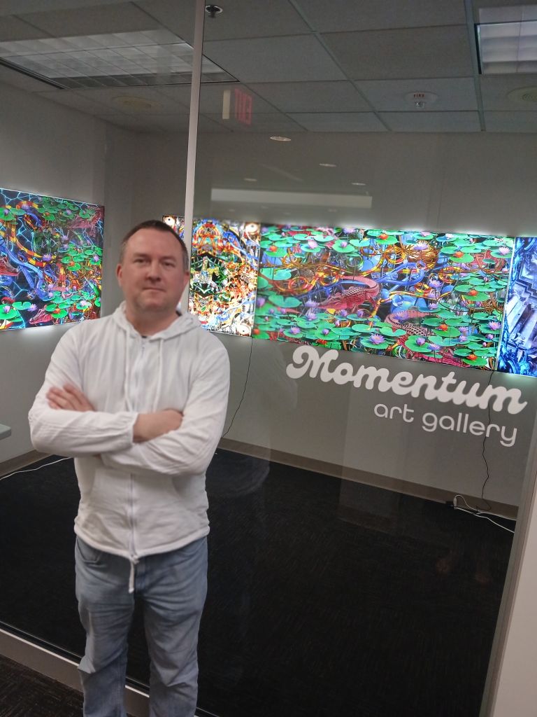 Artist matt corones stands in front of the Momentum West gallery his unique nostalgic, slightly chaotic mixed media art pieces hang on the wall behind him.