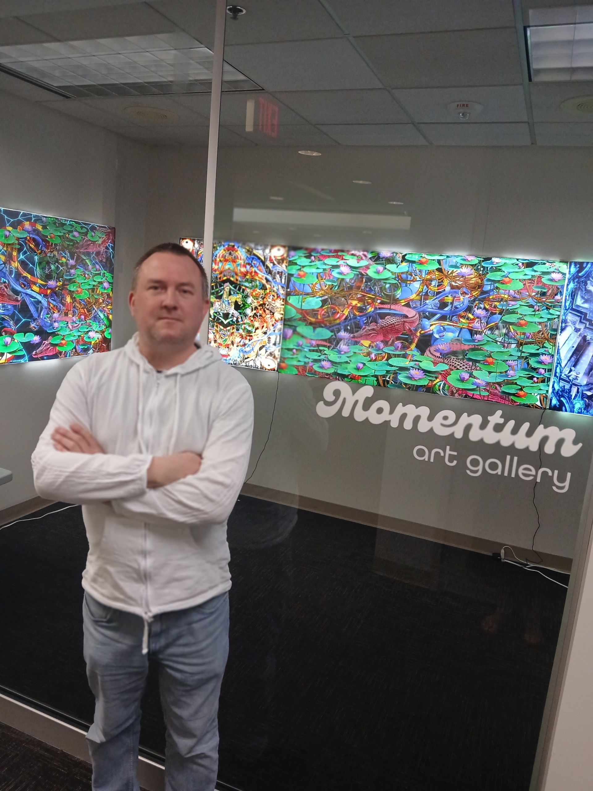 Artist matt corones stands in front of the Momentum West gallery his unique nostalgic, slightly chaotic mixed media art pieces hang on the wall behind him.