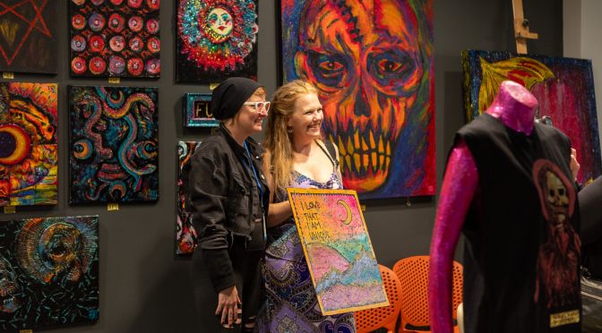 Two women stand for a photo. One is holding a painting. In the background is a gallery of fiery design, including a prominent painting of an orange skull.