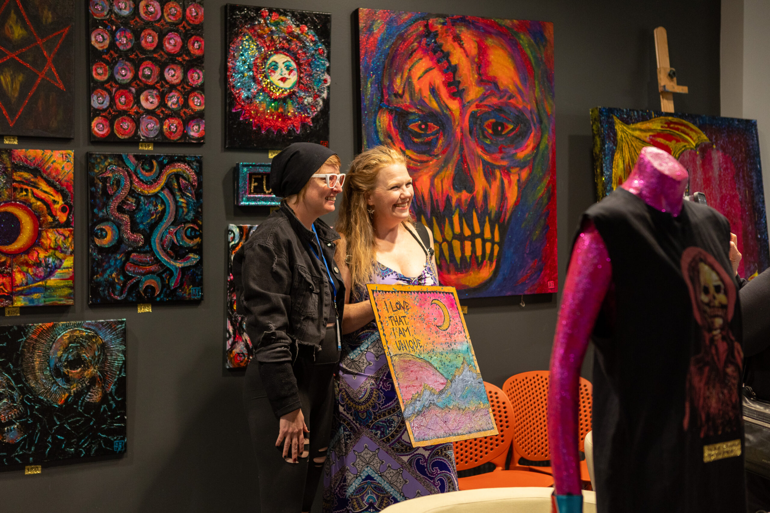 Two women stand for a photo. One is holding a painting. In the background is a gallery of fiery design, including a prominent painting of an orange skull.